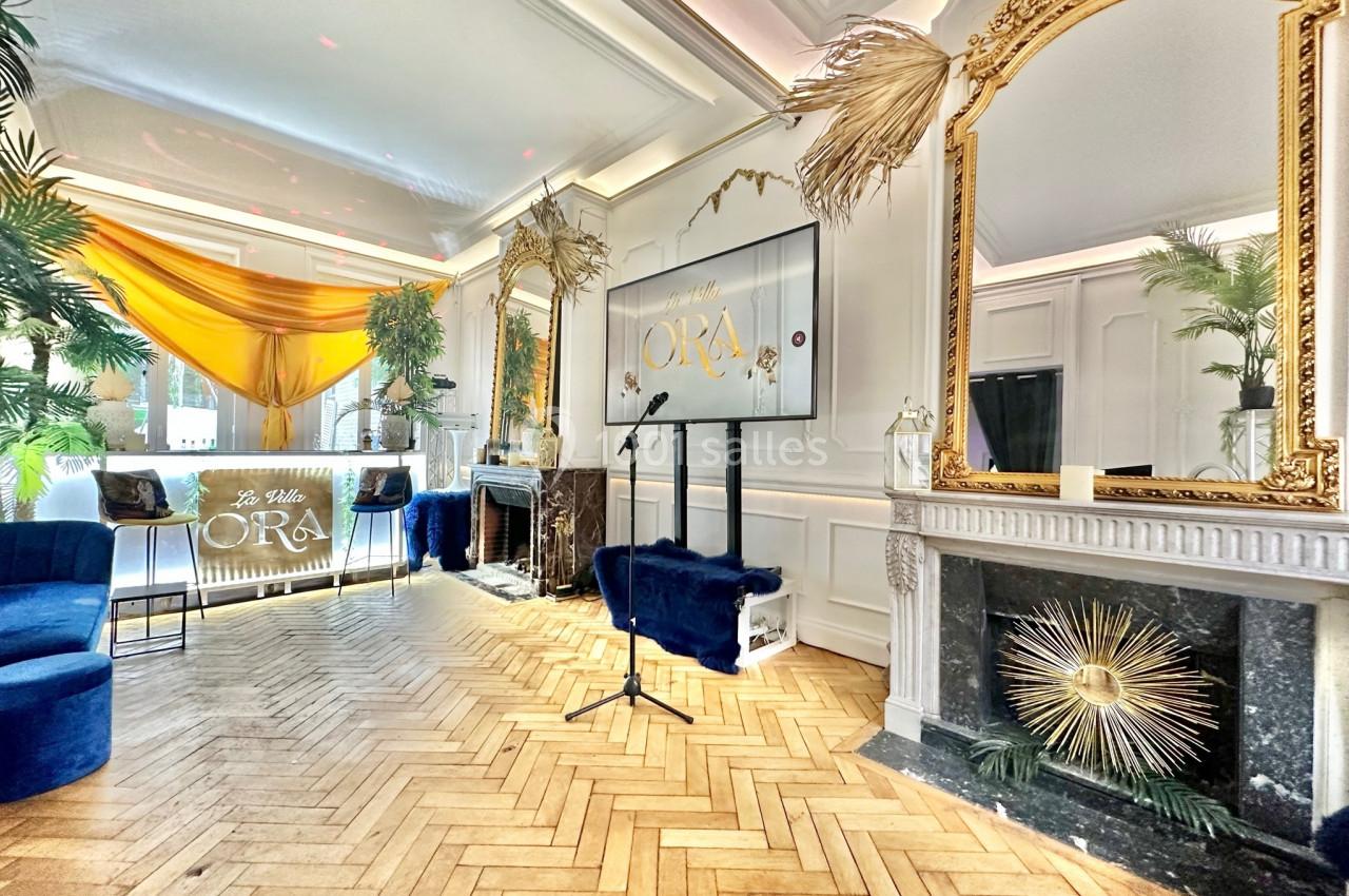 Salle élégante avec parquet en chevrons, miroirs dorés, décoration végétale et écran affichant ’ORA’.