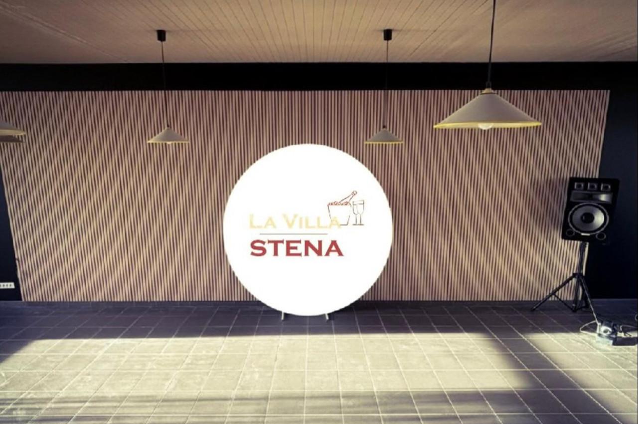 Mur intérieur avec un panneau lumineux circulaire affichant ’La Villa Stena’, entouré de lampes suspendues et d'une enceinte.