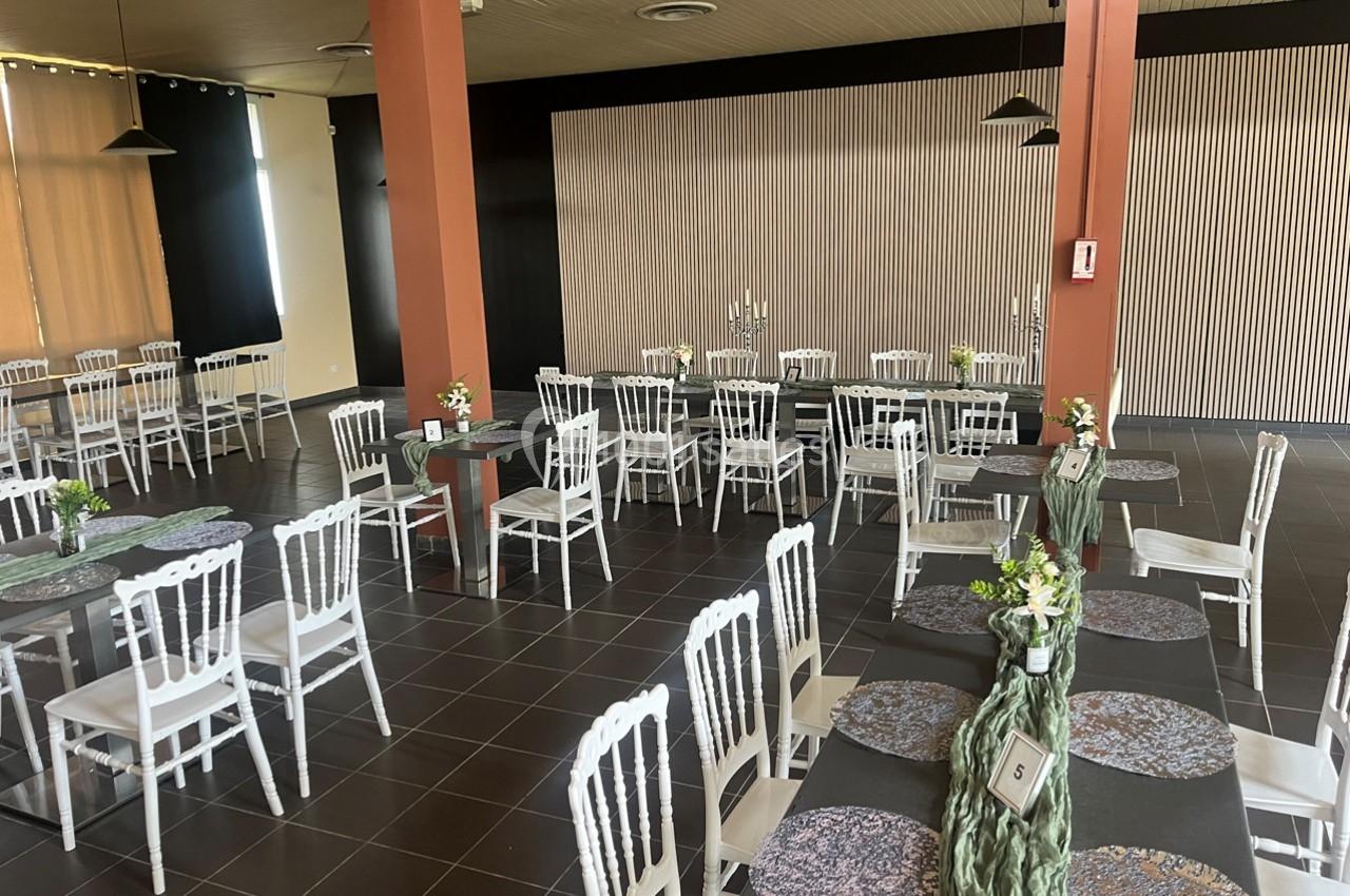 Salle de réception avec des tables dressées, nappes sombres, chaises blanches et décorations florales.