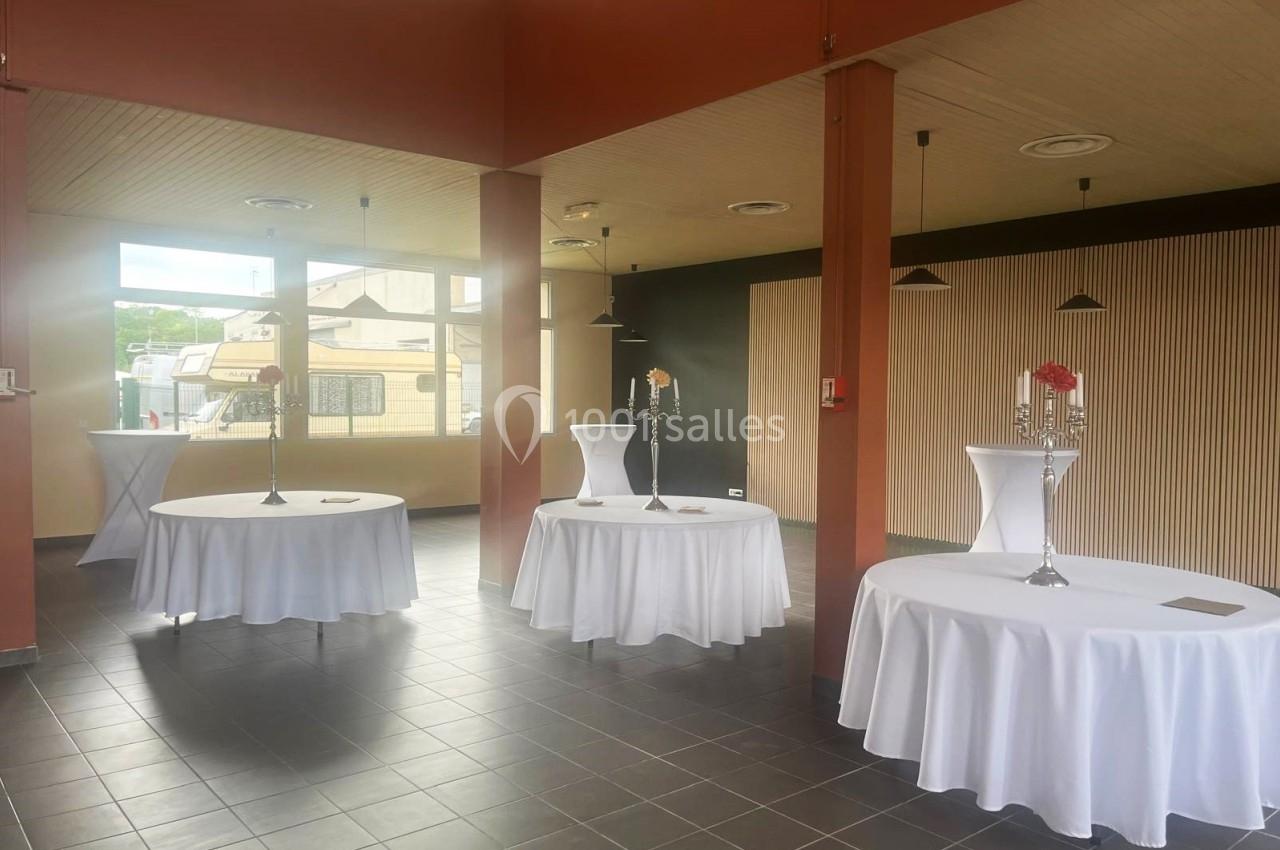Salle lumineuse avec tables rondes recouvertes de nappes blanches, décorées de chandeliers et de fleurs.
