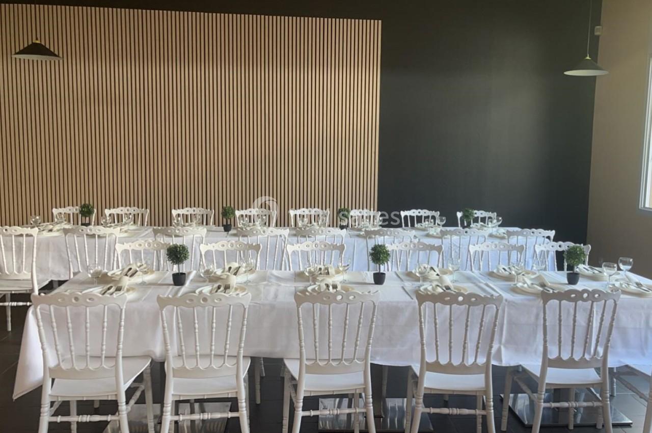 Salle de réception avec des tables dressées, nappes blanches, chaises blanches et petites plantes en centre de table.