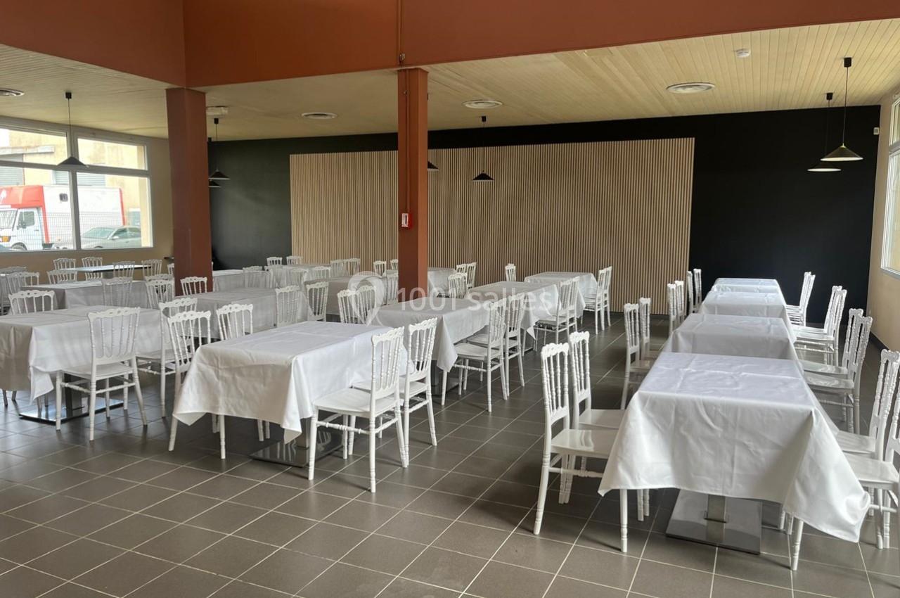 Salle de restaurant avec des tables rectangulaires couvertes de nappes blanches et des chaises blanches alignées.