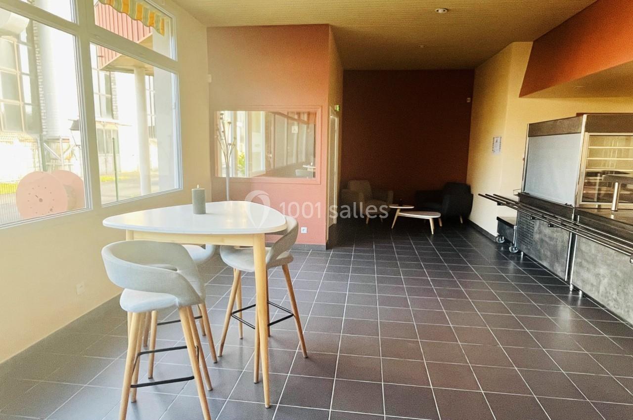 Salle lumineuse avec table haute, chaises, coin salon et grandes fenêtres donnant sur l'extérieur.