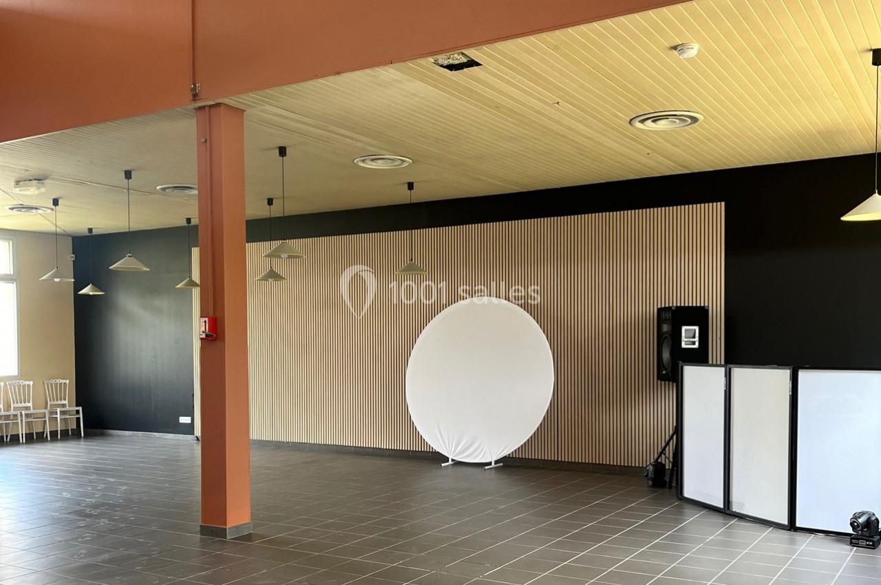 Salle vide avec sol carrelé, murs noirs et bois, cercle blanc en fond, chaises et équipement audio visibles.