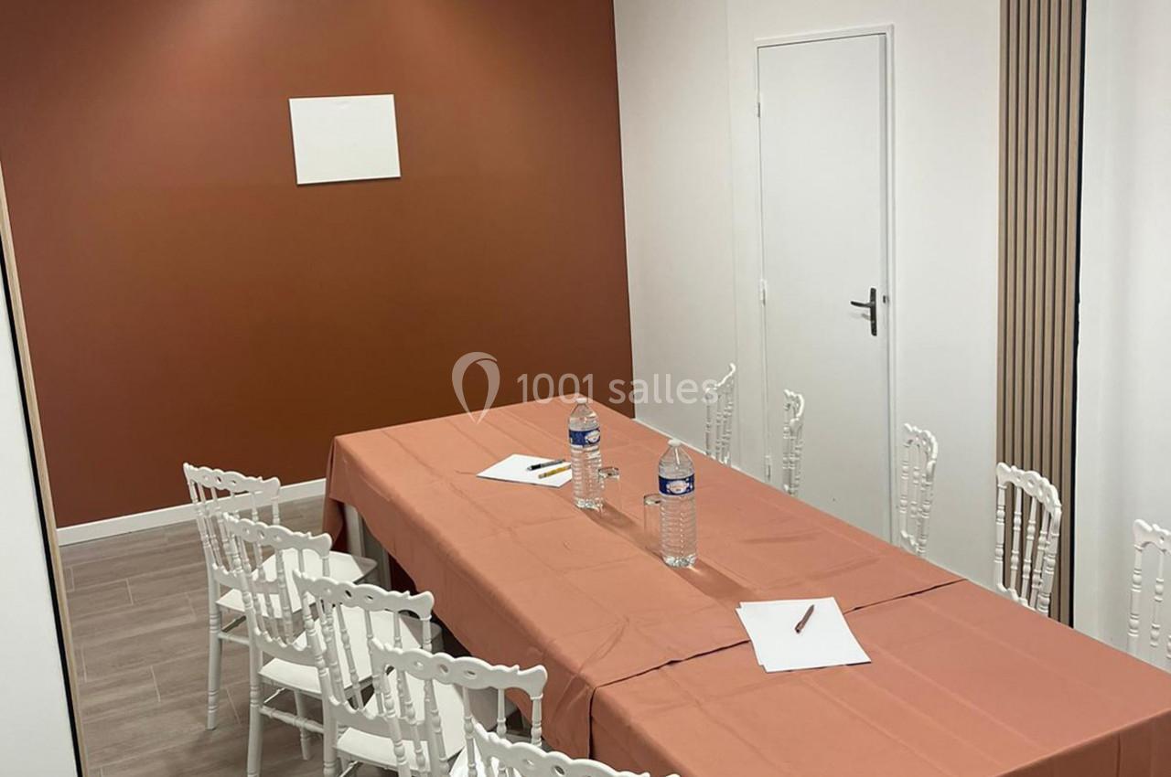 Salle de réunion avec table rectangulaire couverte d'une nappe orange, chaises blanches et fournitures de bureau.
