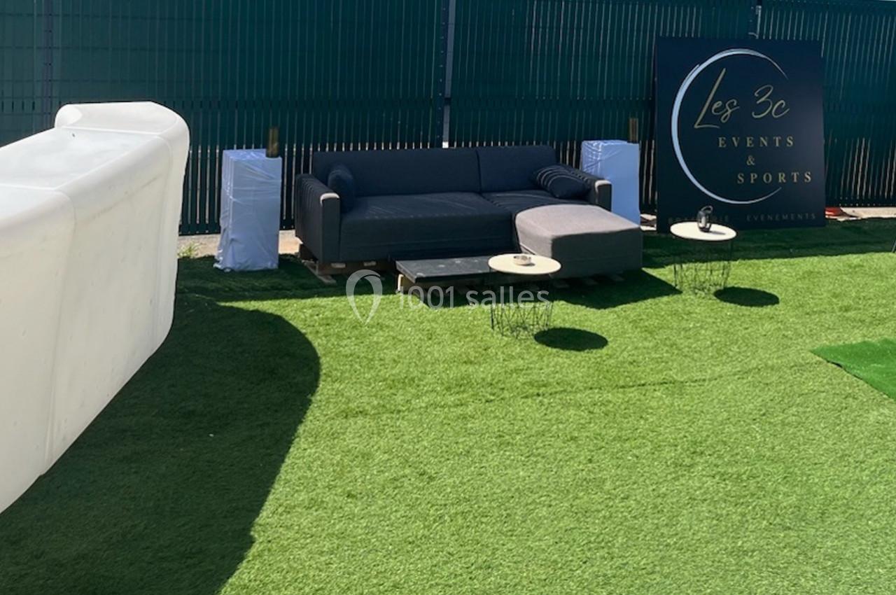 Canapé gris avec table basse sur pelouse synthétique, près d'une enseigne ’Les 3C Events & Sports’.