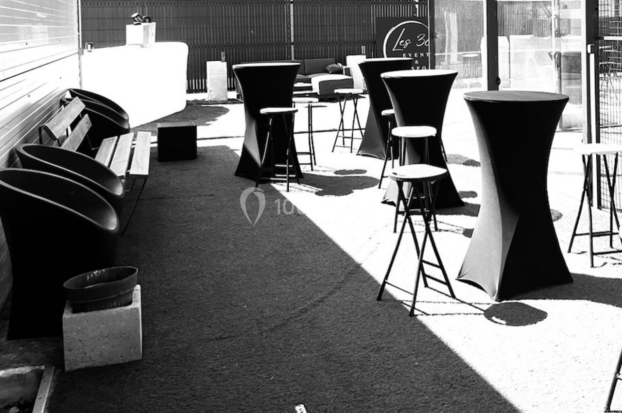 Terrasse aménagée avec tables hautes, tabourets et bancs, éclairée par la lumière naturelle.