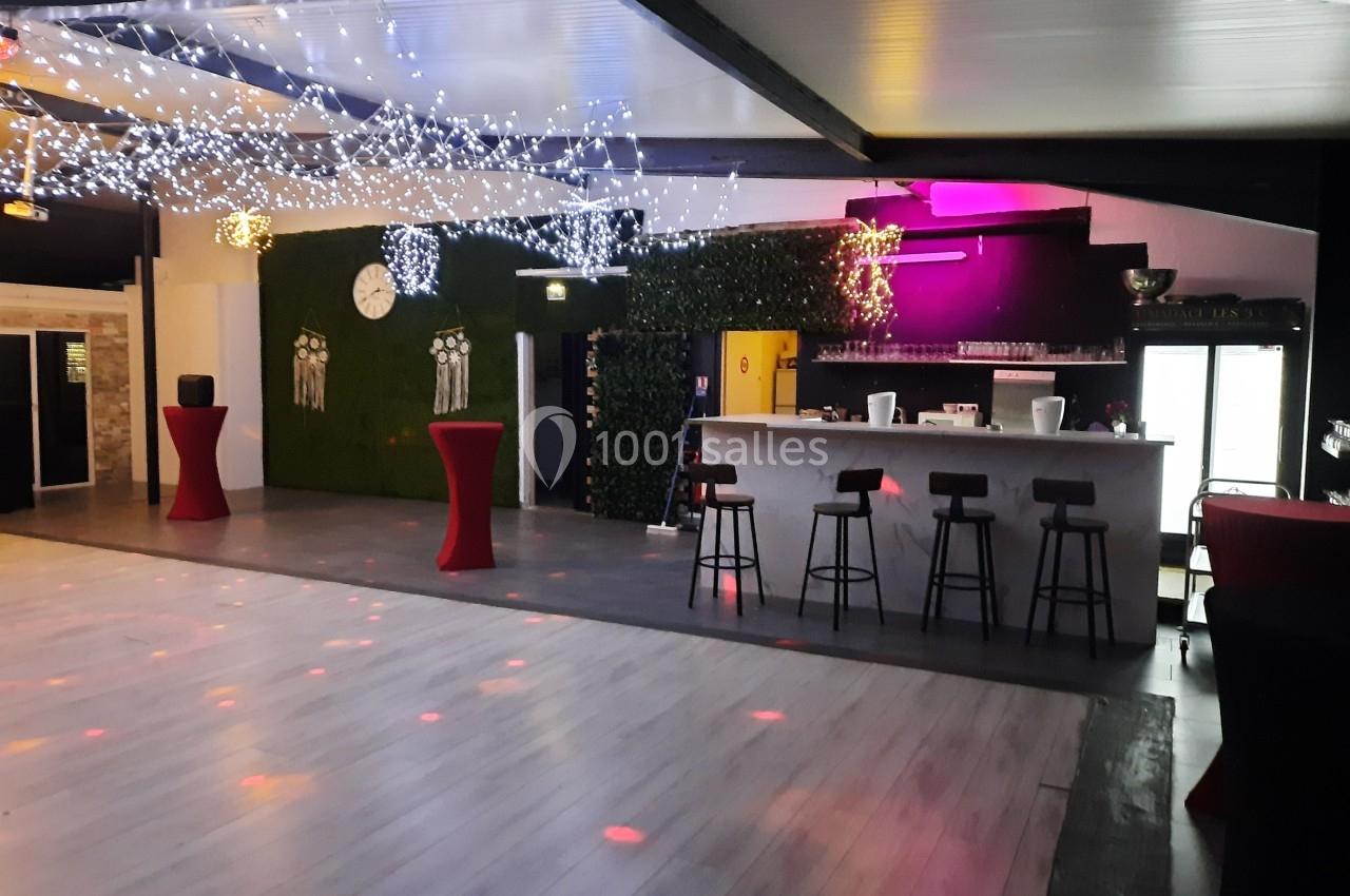 Salle de réception avec piste de danse, bar équipé de tabourets, tables hautes et décorations lumineuses au plafond.