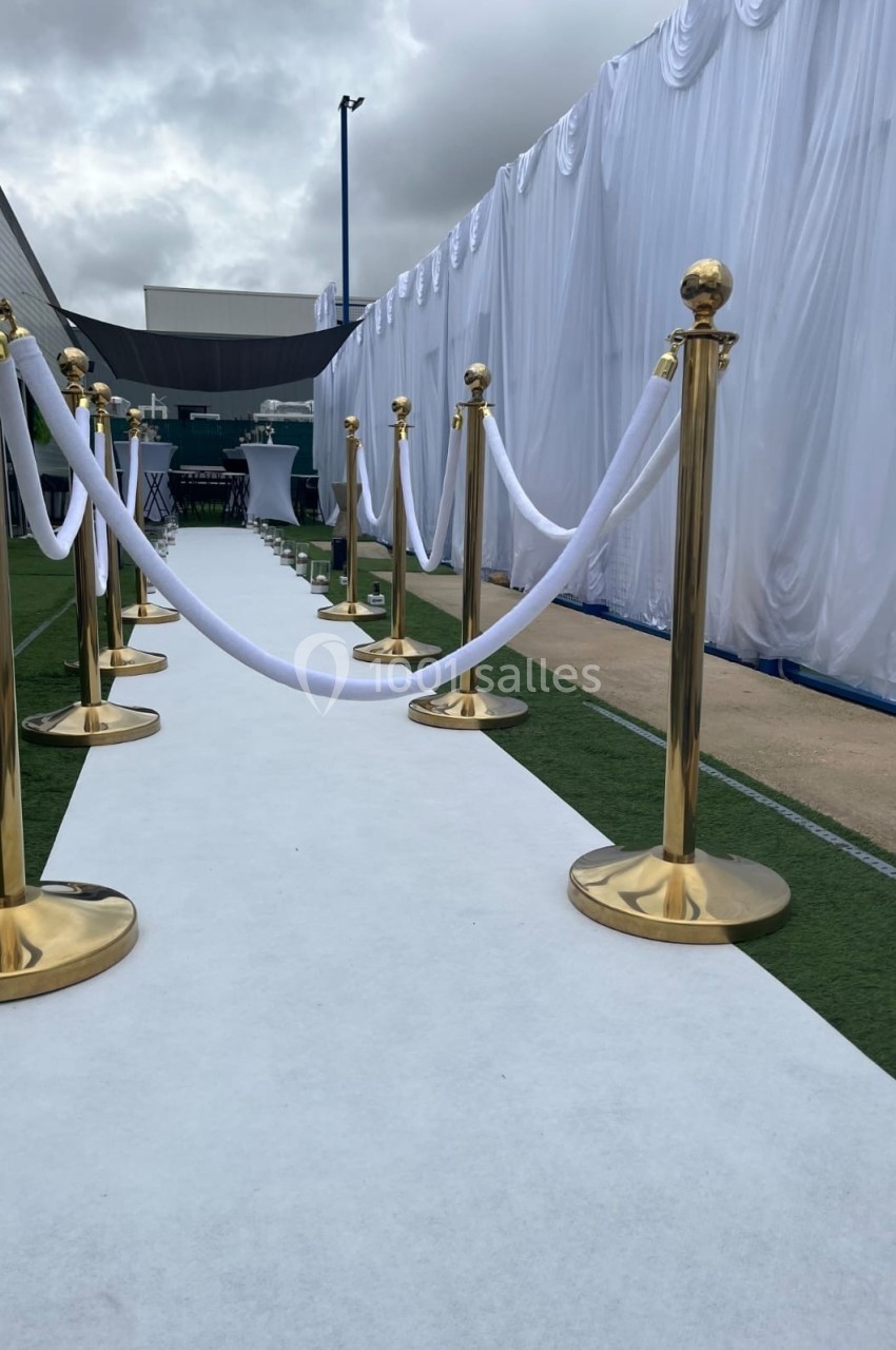 Tapis blanc bordé de poteaux dorés et cordons blancs, menant à une structure décorée sous un ciel nuageux.