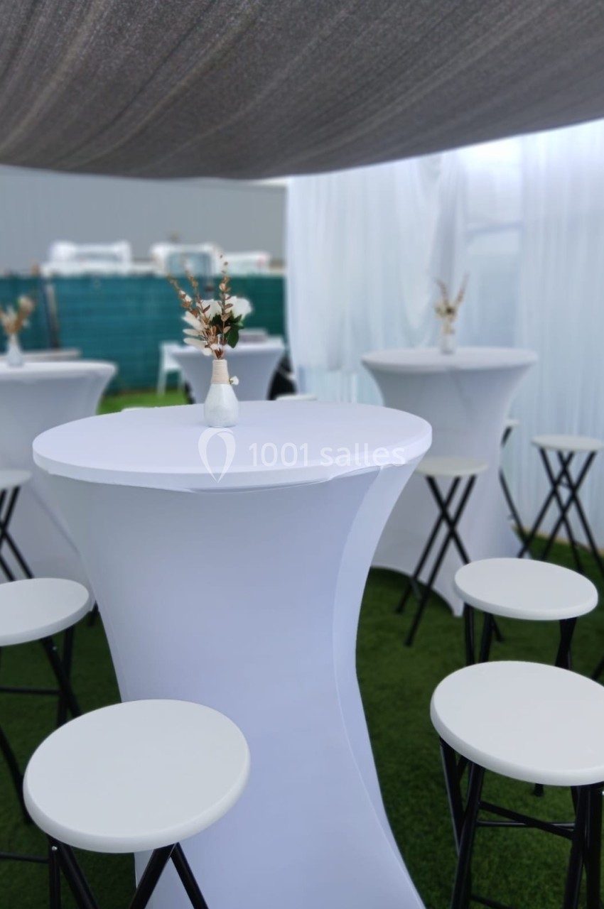 Tables hautes blanches avec tabourets assortis, décorées de petits vases fleuris, dans un espace extérieur couvert.