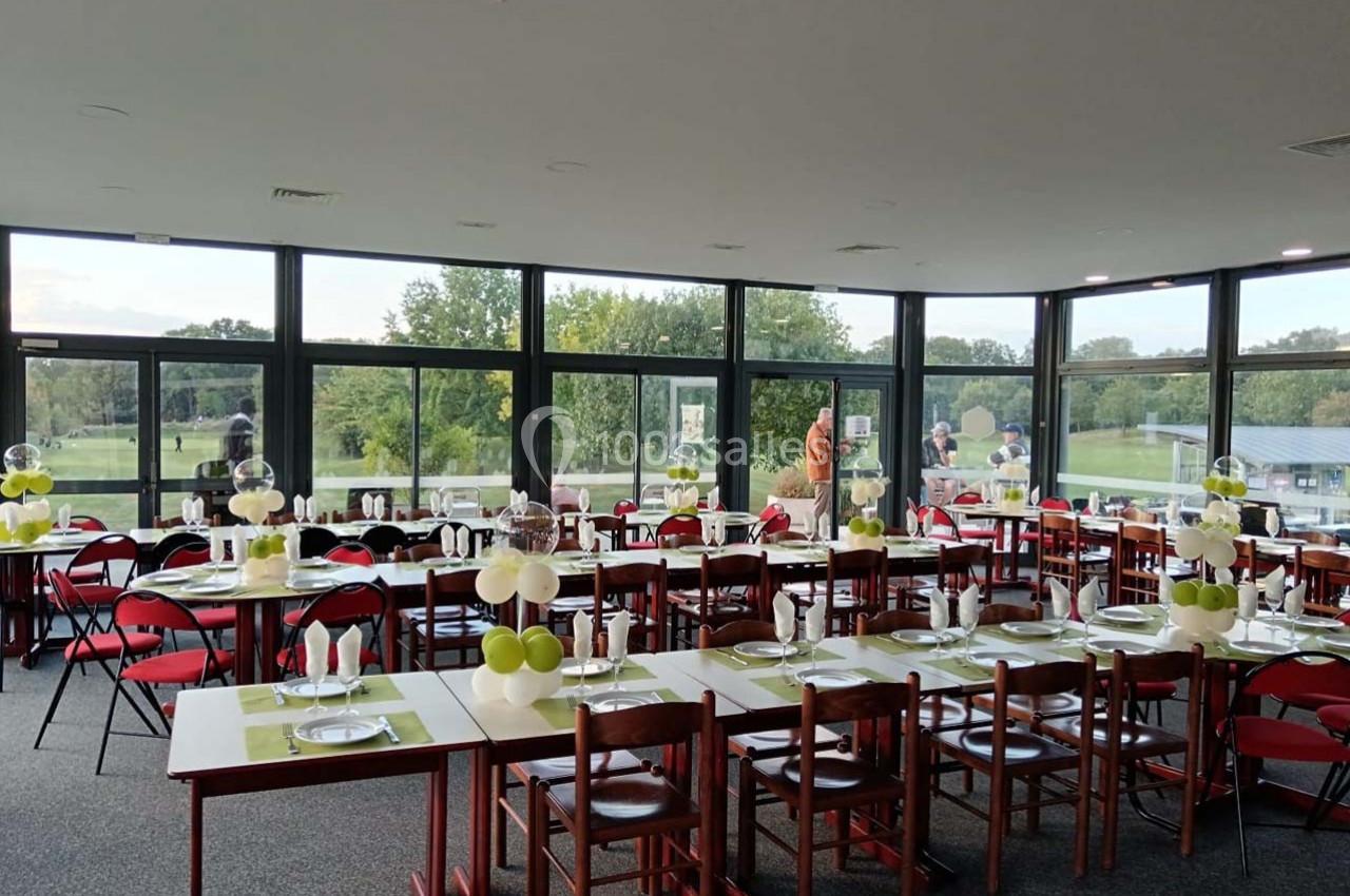 Salle lumineuse avec grandes baies vitrées, tables dressées avec nappes vertes, chaises rouges et décorations de table.