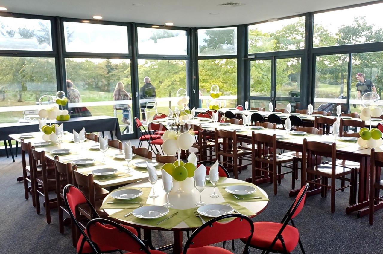 Salle lumineuse avec tables rondes dressées, nappes vertes, décorations de pommes et chaises rouges, vue sur un jardin.