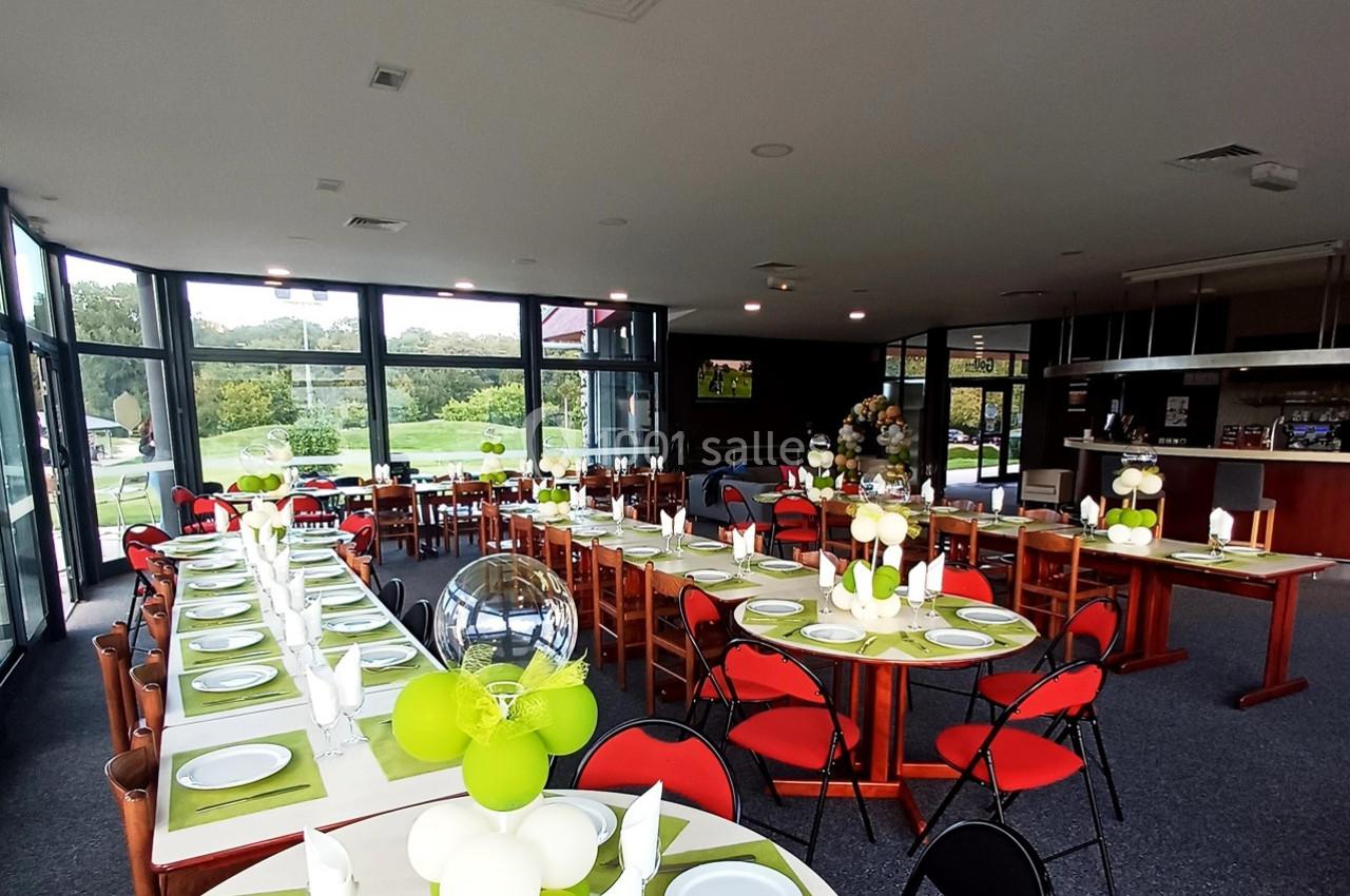Salle de réception lumineuse avec tables dressées, décorations vertes et blanches, et vue sur un jardin à travers de grandes…