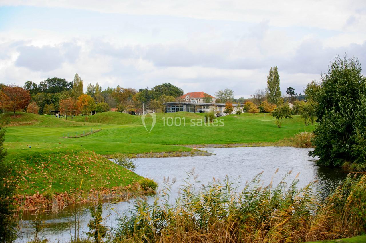 Paysage de golf avec un étang au premier plan, des pelouses verdoyantes et un bâtiment entouré d'arbres en arrière-plan.