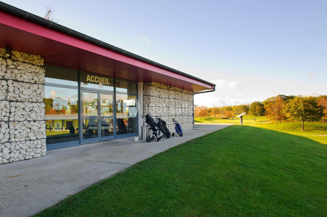 Bâtiment d'accueil avec façade en pierre, devanture vitrée et chariots de golf, entouré d'un terrain verdoyant.