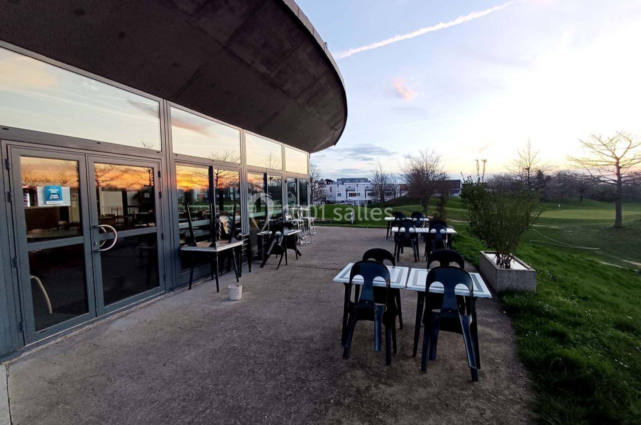 Terrasse extérieure avec tables et chaises, située près d'un bâtiment vitré, entourée d'herbe et d'arbres au coucher du…