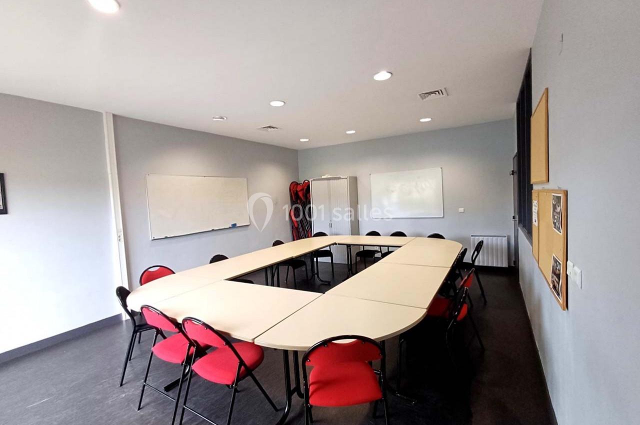 Salle de réunion avec tables disposées en U, chaises rouges, tableau blanc et panneaux d'affichage au mur.