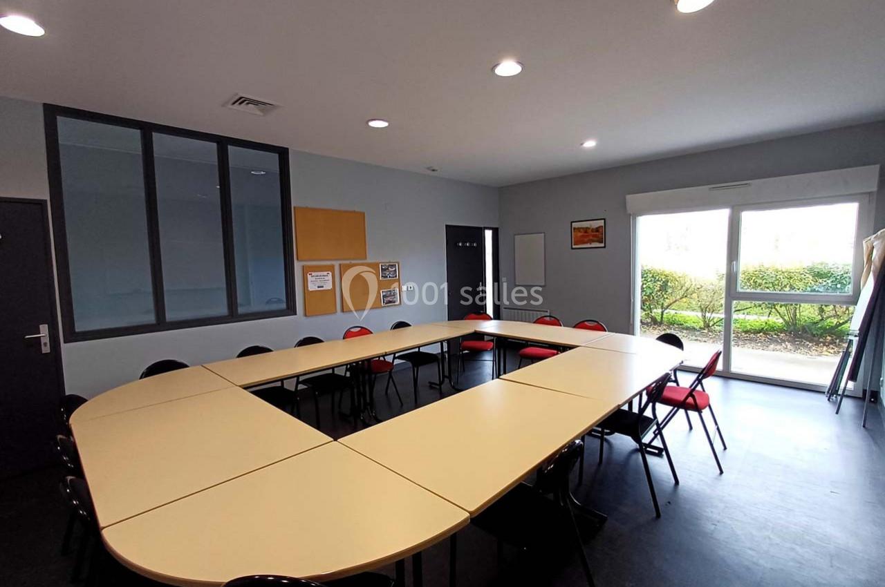 Salle de réunion lumineuse avec tables disposées en U, chaises rouges et noires, et vue sur un espace extérieur verdoyant.