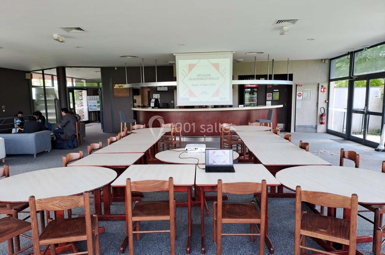 Salle de réunion avec tables disposées en U, chaises en bois, écran de projection allumé et espace d'accueil en arrière-plan.