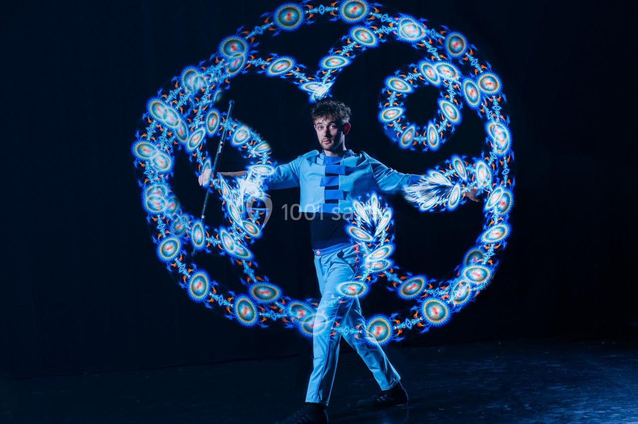 Un homme jongle avec des bâtons lumineux créant des motifs circulaires colorés sur un fond sombre.