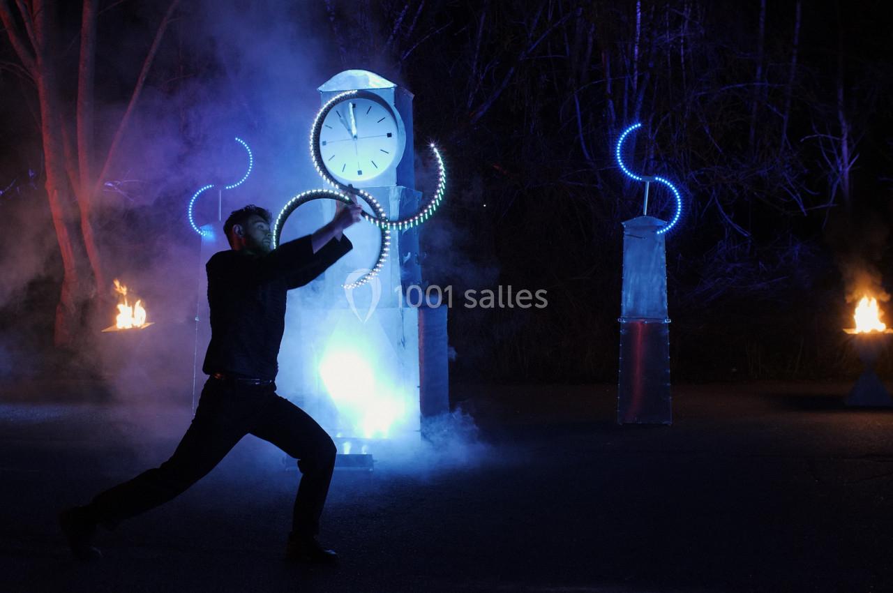 Un homme en mouvement devant une installation lumineuse avec une horloge, entourée de fumée et de flammes.