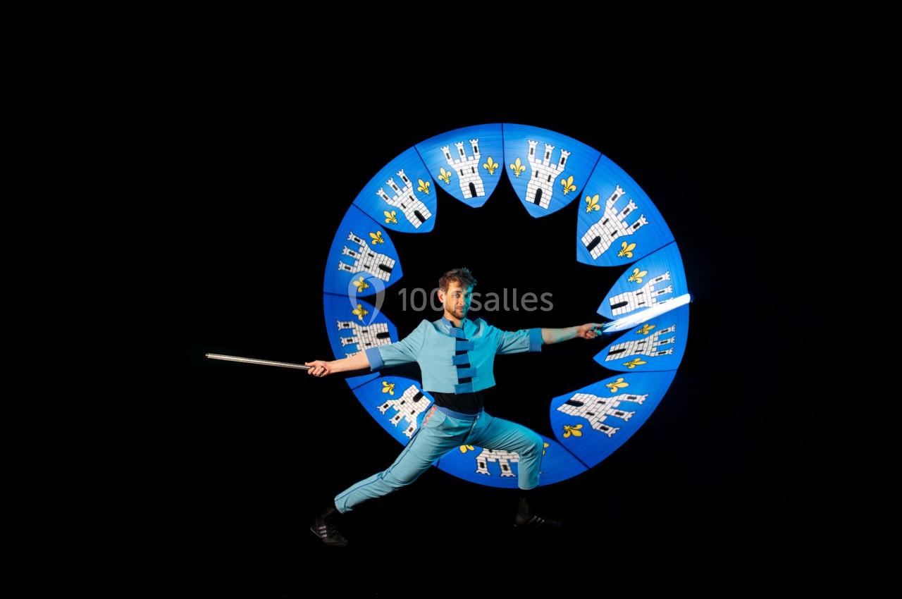 Un artiste en costume bleu manipule un bâton lumineux devant un cercle décoré de châteaux et de fleurs de lys.