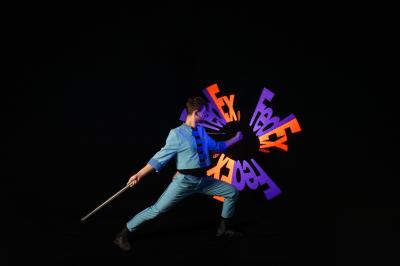 Un jongleur manipule des bâtons rouges dans une galerie lumineuse avec une verrière en arrière-plan.