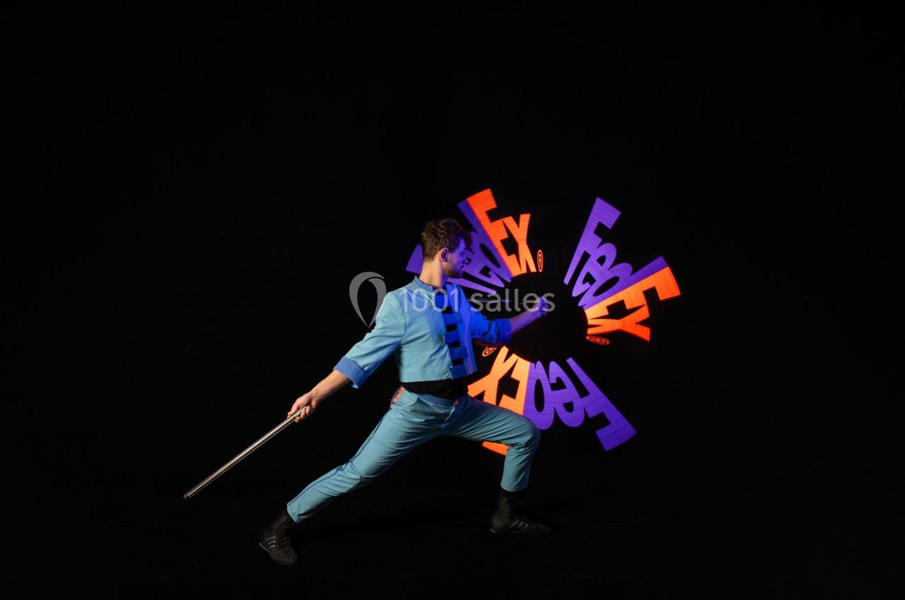 Un artiste en costume bleu manipule un bâton lumineux créant des motifs colorés sur un fond noir.