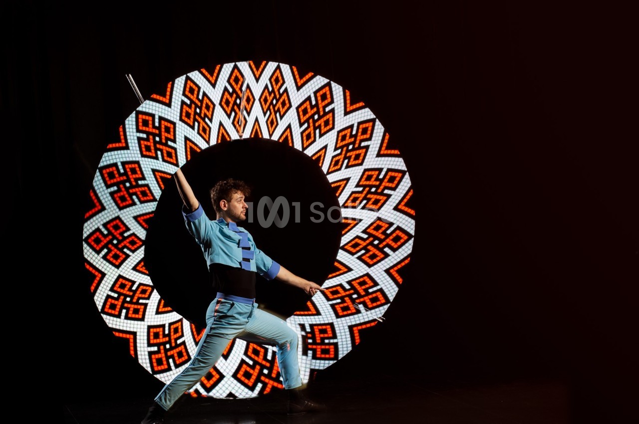 Un homme en tenue bleue effectue une pose dynamique devant un cercle lumineux aux motifs géométriques colorés.