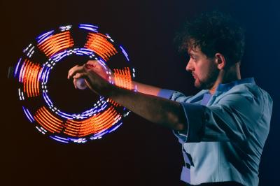 Un jongleur réalise une performance avec des bâtons rouges dans une galerie lumineuse bordée de colonnes.