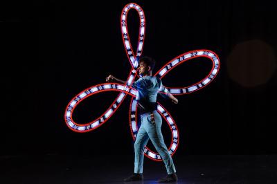 Un jongleur réalise une performance avec des bâtons rouges dans une galerie lumineuse bordée de colonnes.