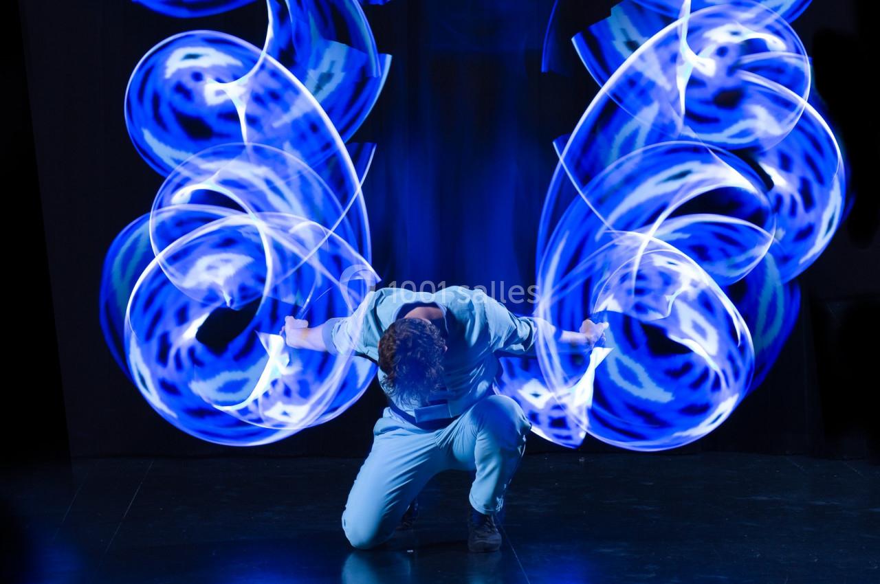 Personne en mouvement, agenouillée, entourée de traînées lumineuses bleues formant des motifs dynamiques.