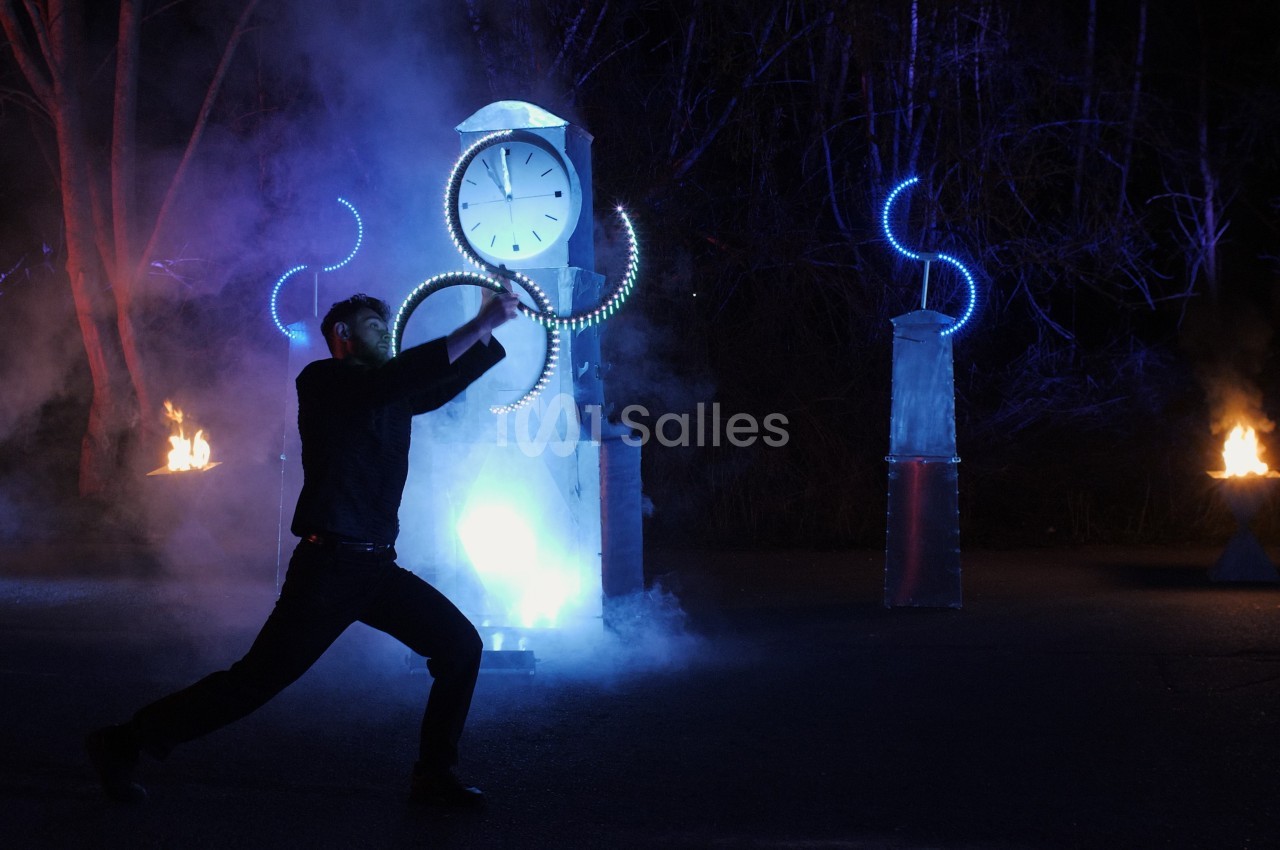 Un homme danse devant une horloge illuminée et des installations lumineuses, entouré de fumée et de flammes.
