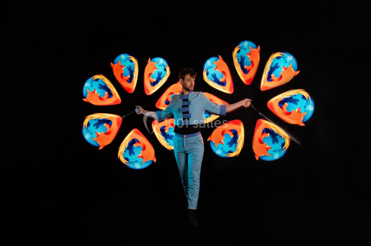 Un jongleur manipule des bâtons lumineux créant des motifs colorés symétriques sur fond noir.