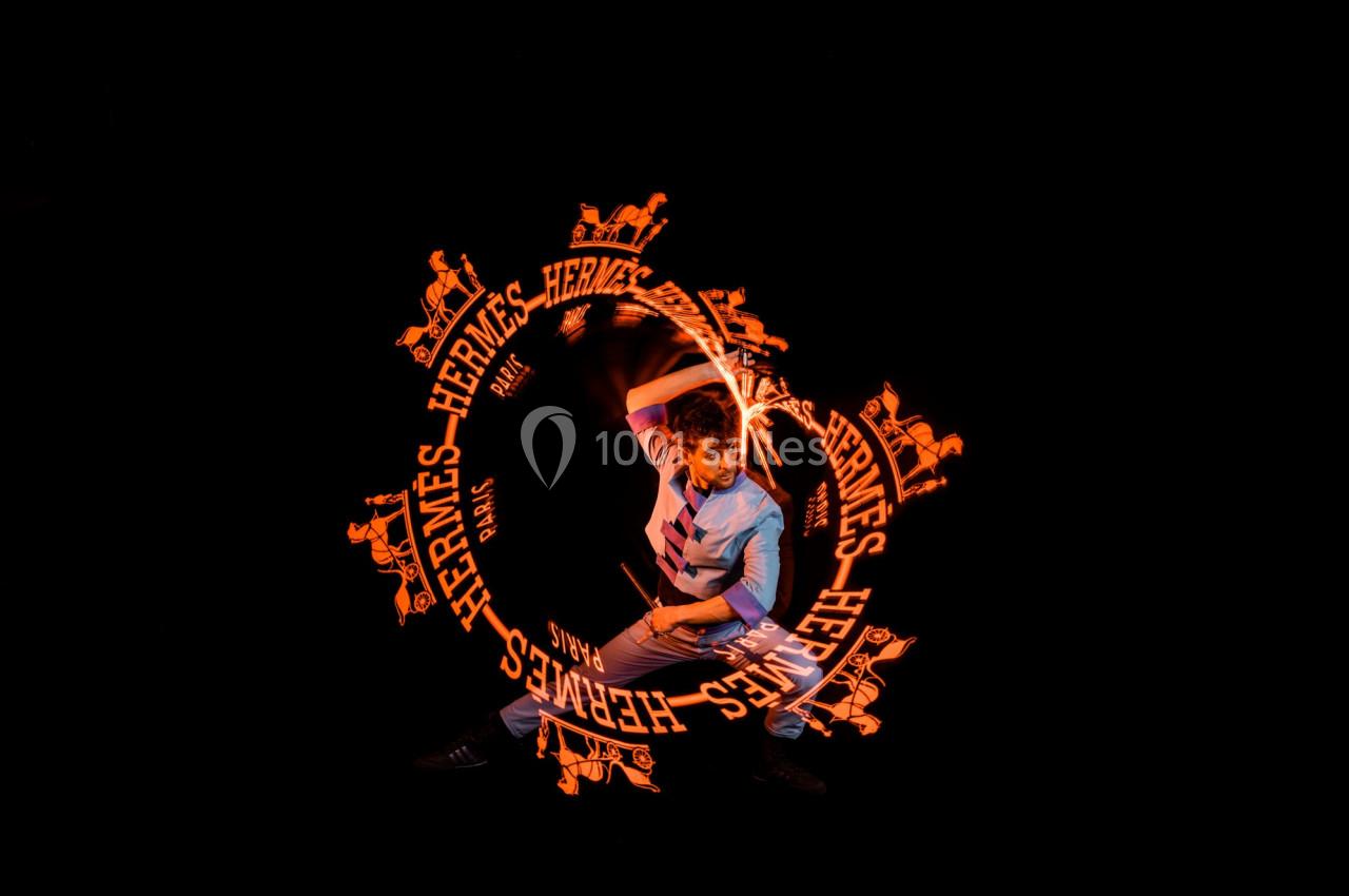 Un homme effectue une performance lumineuse avec des motifs orange et le logo Hermès dans un environnement sombre.