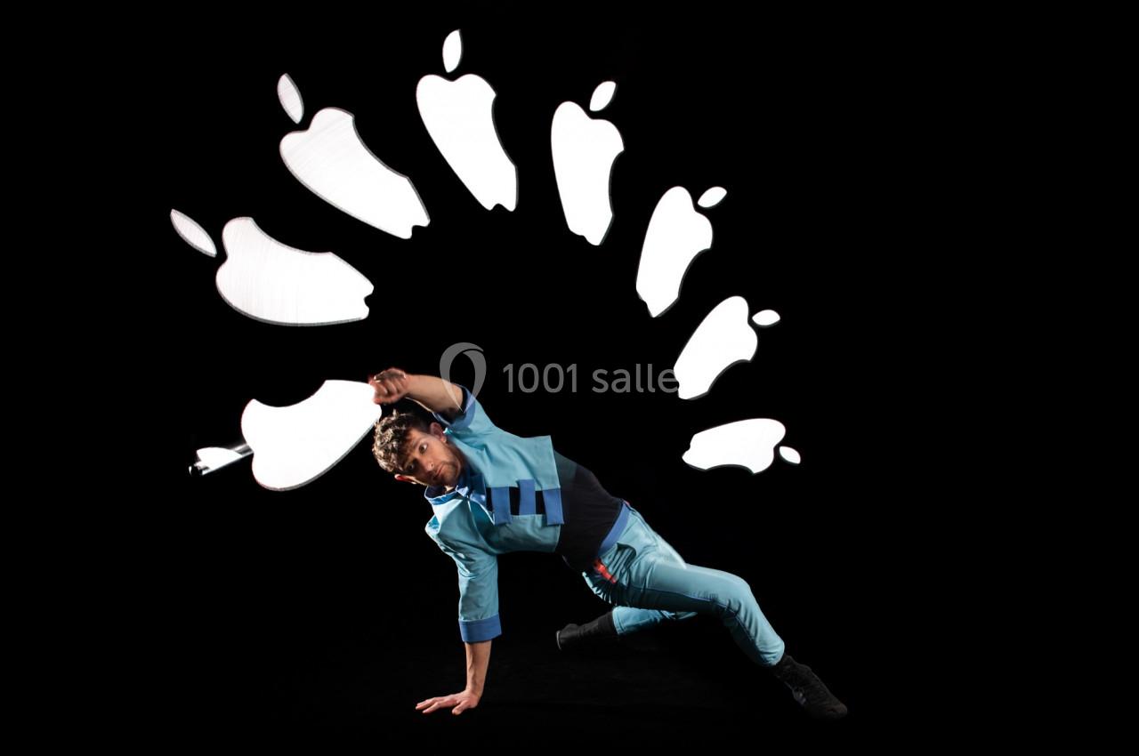 Un danseur en tenue bleue effectue une figure au sol avec des formes lumineuses en arrière-plan sur fond noir.