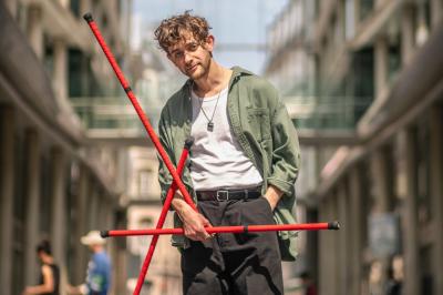 Un jongleur réalise une performance avec des bâtons rouges dans une galerie lumineuse bordée de colonnes.