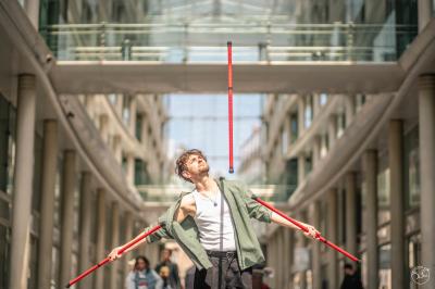Un jongleur réalise une performance avec des bâtons rouges dans une galerie lumineuse bordée de colonnes.