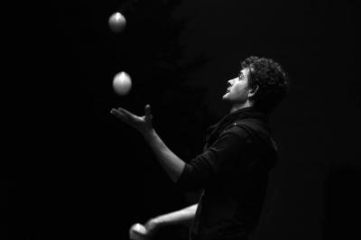 Un jongleur réalise une performance avec des bâtons rouges dans une galerie lumineuse bordée de colonnes.