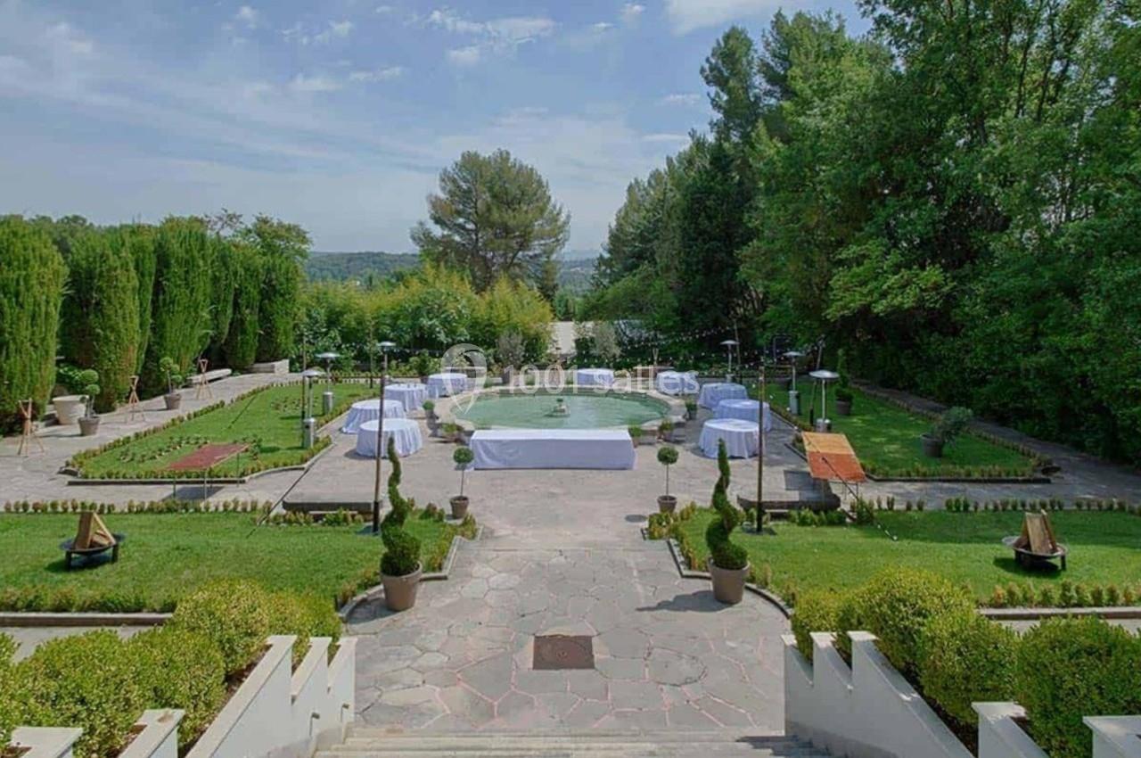 Vue d'un jardin aménagé avec une fontaine centrale, entouré de verdure et de tables décorées pour un événement.