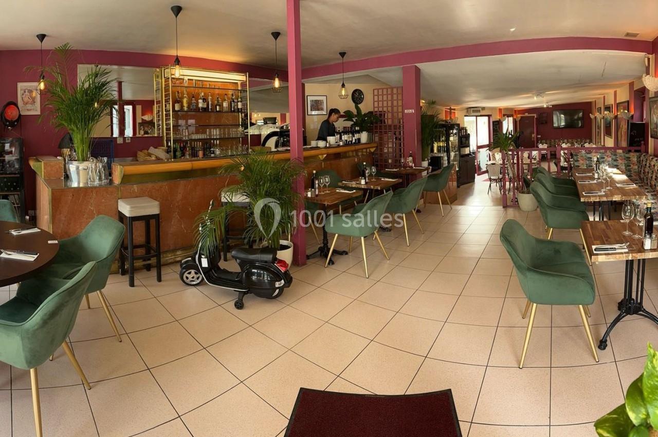 Salle de restaurant lumineuse avec tables en bois, chaises vertes, décorations végétales et comptoir en arrière-plan.