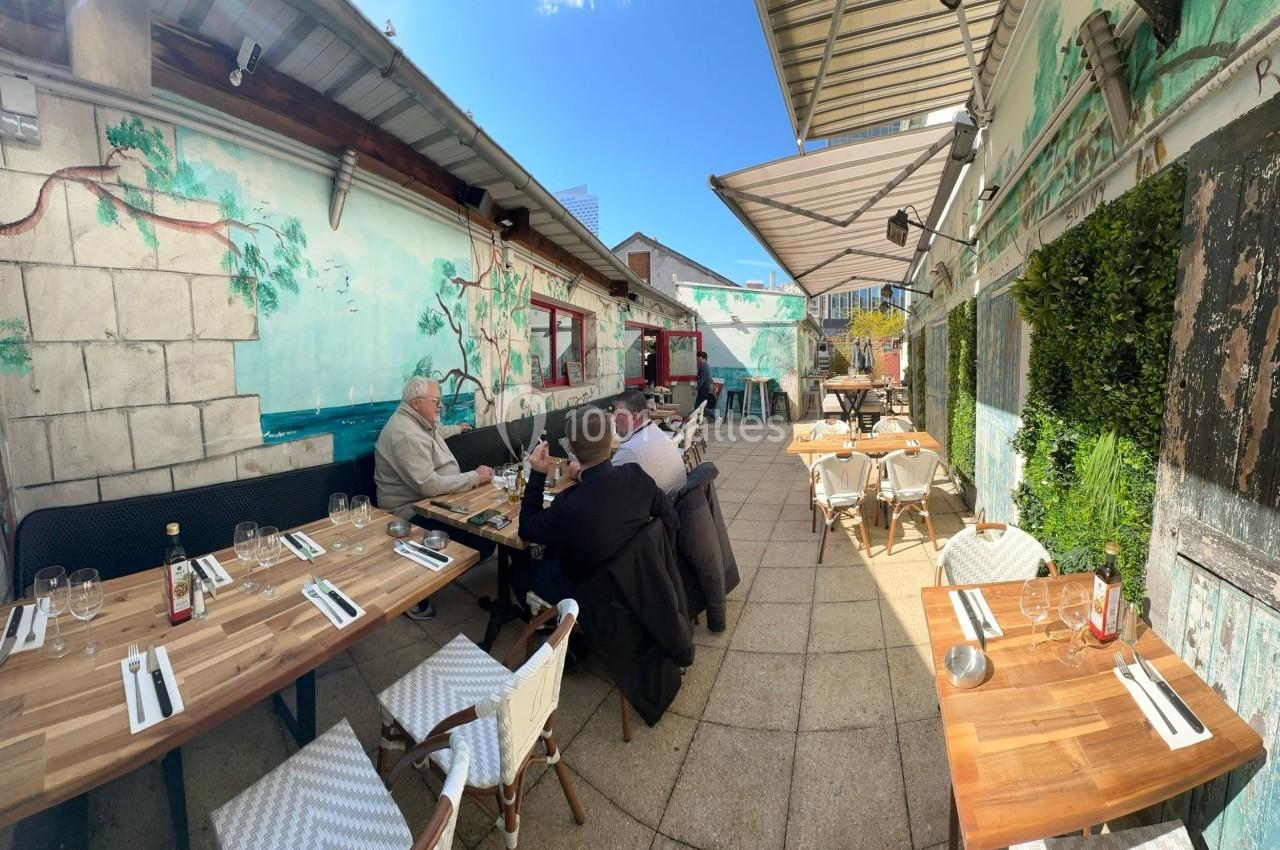 Terrasse extérieure d'un restaurant avec des tables en bois, des chaises blanches et des clients attablés sous un ciel…