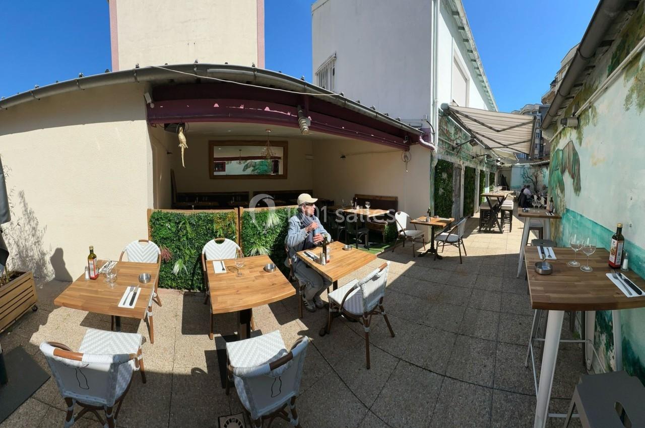 Terrasse extérieure d'un restaurant avec tables en bois, chaises blanches et un homme assis sous un ciel dégagé.