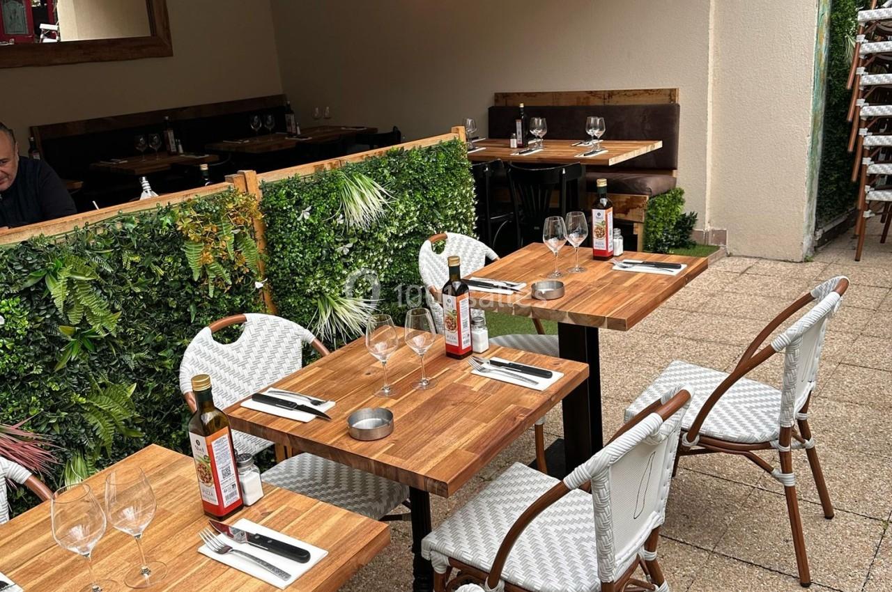 Terrasse d'un restaurant avec tables en bois, chaises blanches, bouteilles d'huile et verdure en décoration.