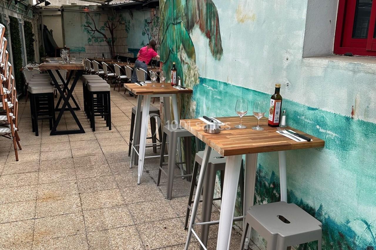 Terrasse extérieure avec tables hautes en bois, chaises, couverts disposés et une personne en arrière-plan.