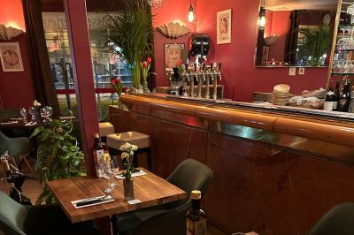 Salle de restaurant lumineuse avec tables dressées, chaises vertes, bar en bois et décoration végétale.