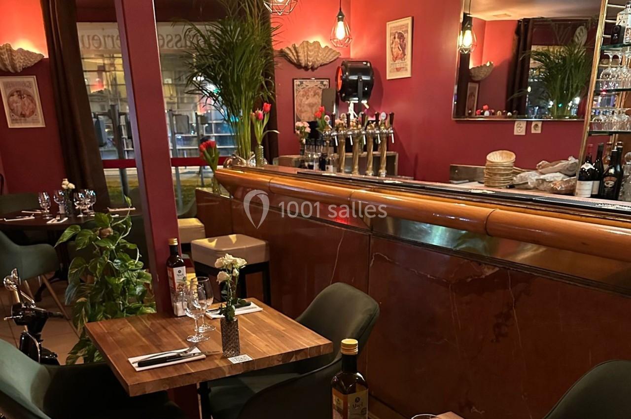 Salle de restaurant chaleureuse avec tables dressées, bar en bois, décor rouge et éclairage tamisé.