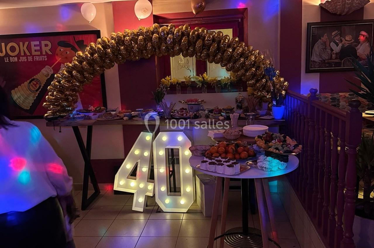 Décoration festive avec arche de ballons dorés, chiffre lumineux ’40’ et buffet varié dans une salle éclairée.