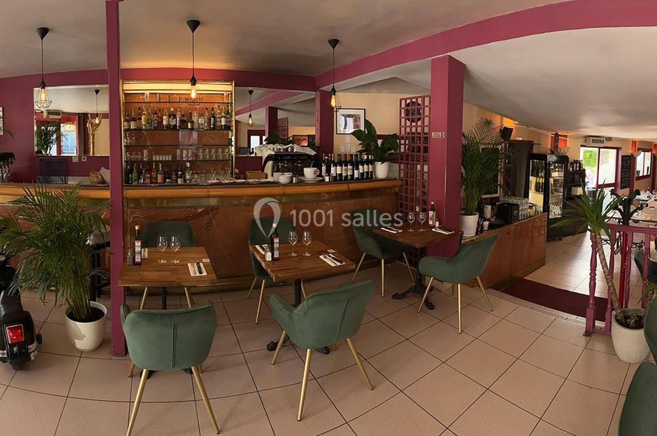Salle de restaurant lumineuse avec tables dressées, chaises vertes, bar en bois et décoration végétale.