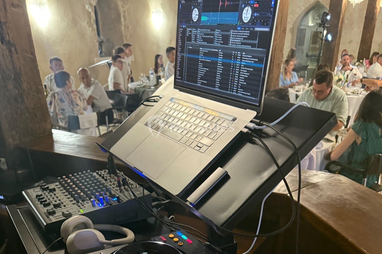 Poste de DJ avec ordinateur portable, table de mixage et casque, dans une salle de réception avec des invités en arrière…
